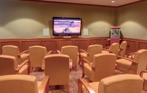 A small theater room with several rows of cushioned armchairs facing a wall-mounted flat screen TV. The room has warm lighting, wood paneling on the lower half of the walls, and a popcorn machine in the corner.