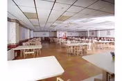 Large dining room with multiple tables and chairs arranged in rows. The room has a tiled ceiling with fluorescent lighting, patterned wallpaper, and a linoleum floor with a checkered pattern. There are windows along the left wall and a few framed pictures on the walls.