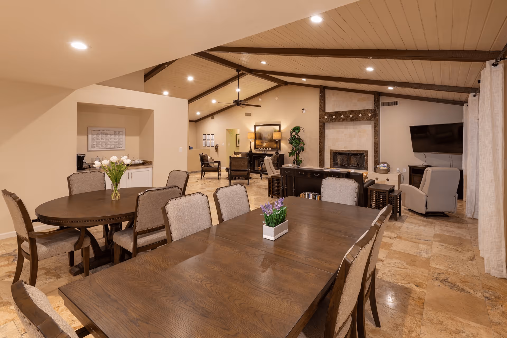 Interior view of a senior living facility common area with two wooden dining tables surrounded by upholstered chairs, a fireplace with a tiled surround, a wall-mounted TV, and comfortable seating including armchairs. The room has a high ceiling with exposed beams and recessed lighting, and the floor is covered with large beige tiles.