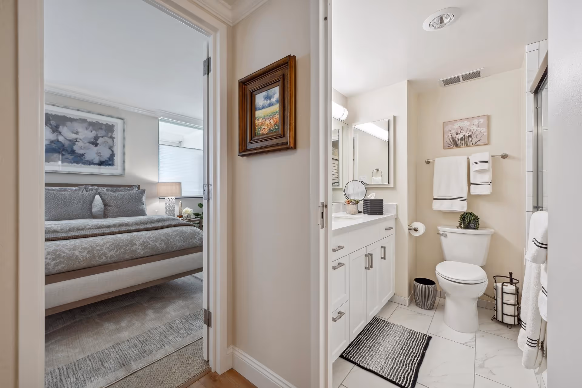 View of a bedroom and an adjacent bathroom in a senior living facility. The bedroom features a neatly made bed with gray patterned bedding, a bedside lamp, and a large window with blinds. The bathroom has a white vanity with a mirror, a toilet with a small plant on top, white towels hanging on racks, a striped black and white rug on the floor, and a shower area partially visible.