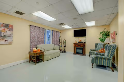 A cozy living room with a beige sofa, two striped armchairs, a wooden side table with pumpkins, a flat-screen TV mounted above a fireplace, and a potted plant in the corner. The room has light yellow walls, patterned curtains on the window, and fluorescent ceiling lights.