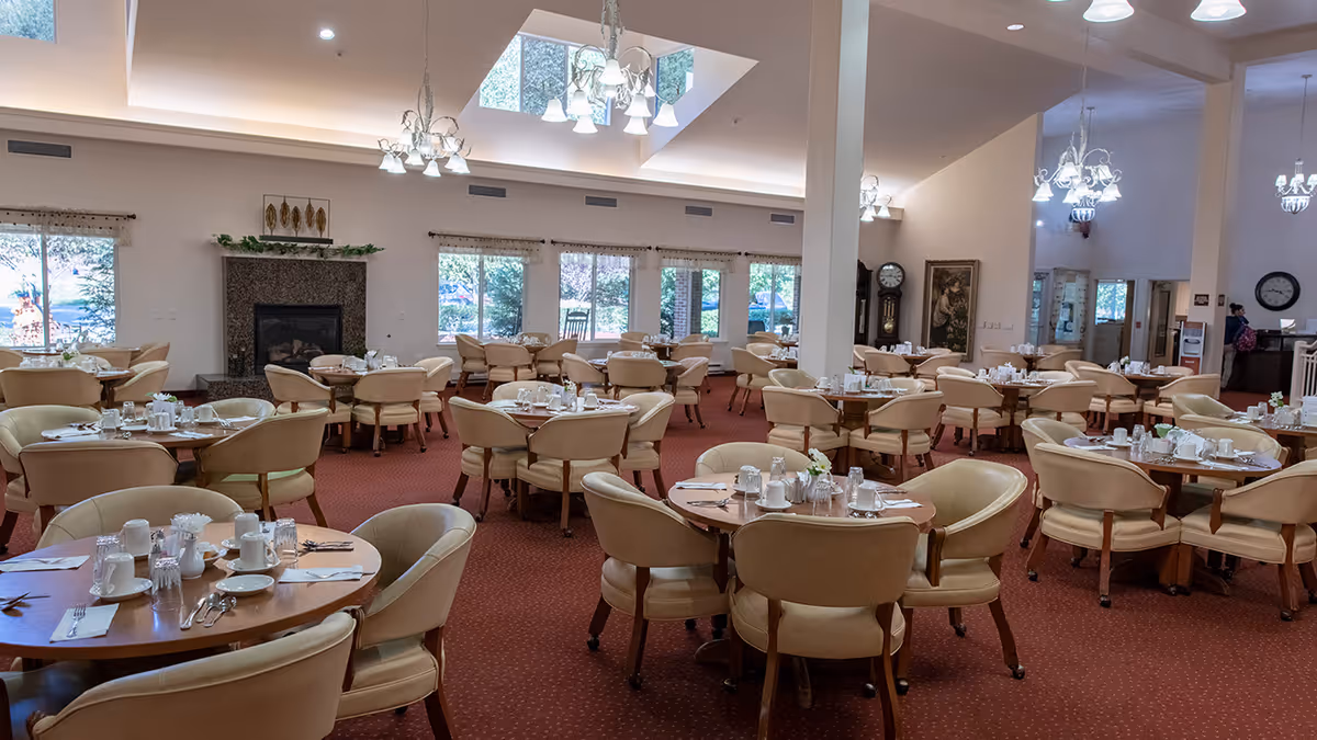 A spacious dining room with multiple round wooden tables each surrounded by beige cushioned chairs. The tables are set with white cups, plates, silverware, and glassware. The room features large windows letting in natural light, a fireplace with decorative greenery above it, and several chandeliers hanging from the ceiling. The carpet is red, and there are paintings and a grandfather clock along the walls.