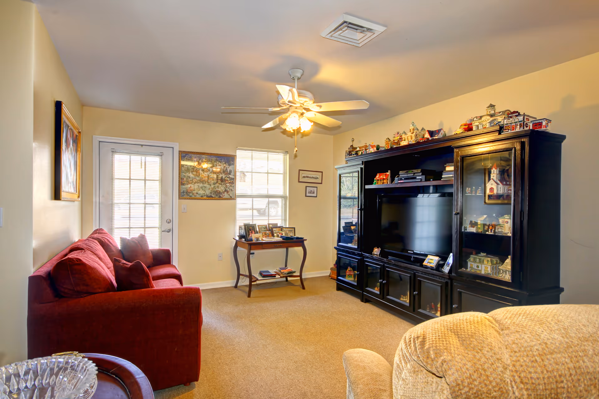 A cozy living room with a red sofa on the left, a beige armchair in the foreground, and a large black entertainment center on the right holding a flat-screen TV and various decorative items. There is a ceiling fan with lights above, a small table with framed photos near a window and a door with blinds, and light yellow walls.