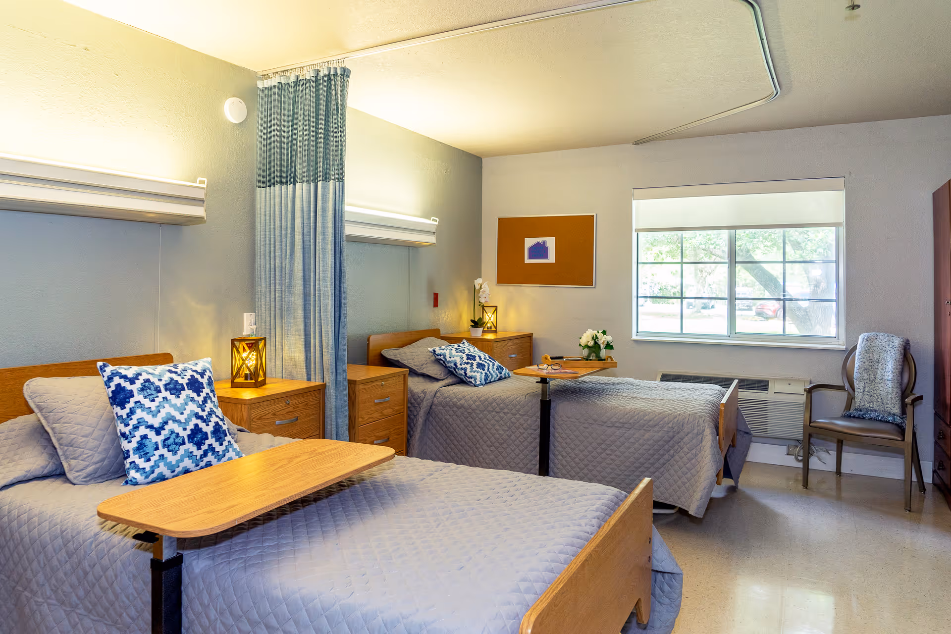 A nursing home room with two single beds, each with gray quilted bedding and a blue patterned pillow. Between the beds is a wooden nightstand with a decorative lantern. A blue privacy curtain hangs between the beds. There is a window with a white blind, a chair with a folded blanket, and a wooden cabinet. Each bed has an adjustable overbed table, and the room is softly lit with wall-mounted lights above each bed.