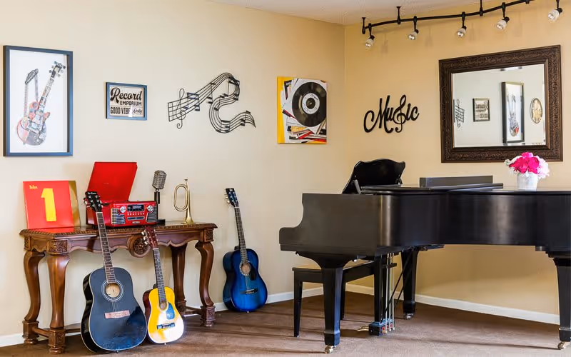 A music-themed room featuring a black grand piano with a vase of pink flowers on top, three acoustic guitars leaning against a wooden table, a red record player on the table, and wall decorations including framed pictures of guitars, musical notes, a vinyl record, and the word 'Music'. A large ornate mirror hangs on the wall reflecting part of the room.
