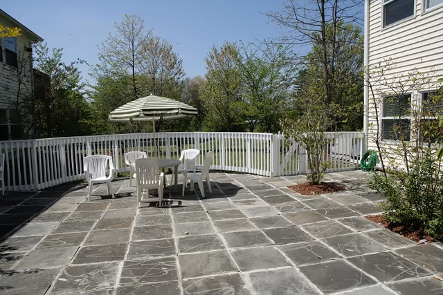 Outdoor patio area with a stone tile floor, a round table with a striped umbrella, and four white plastic chairs. The patio is enclosed by a white fence, with trees and greenery visible beyond the fence under a clear blue sky.