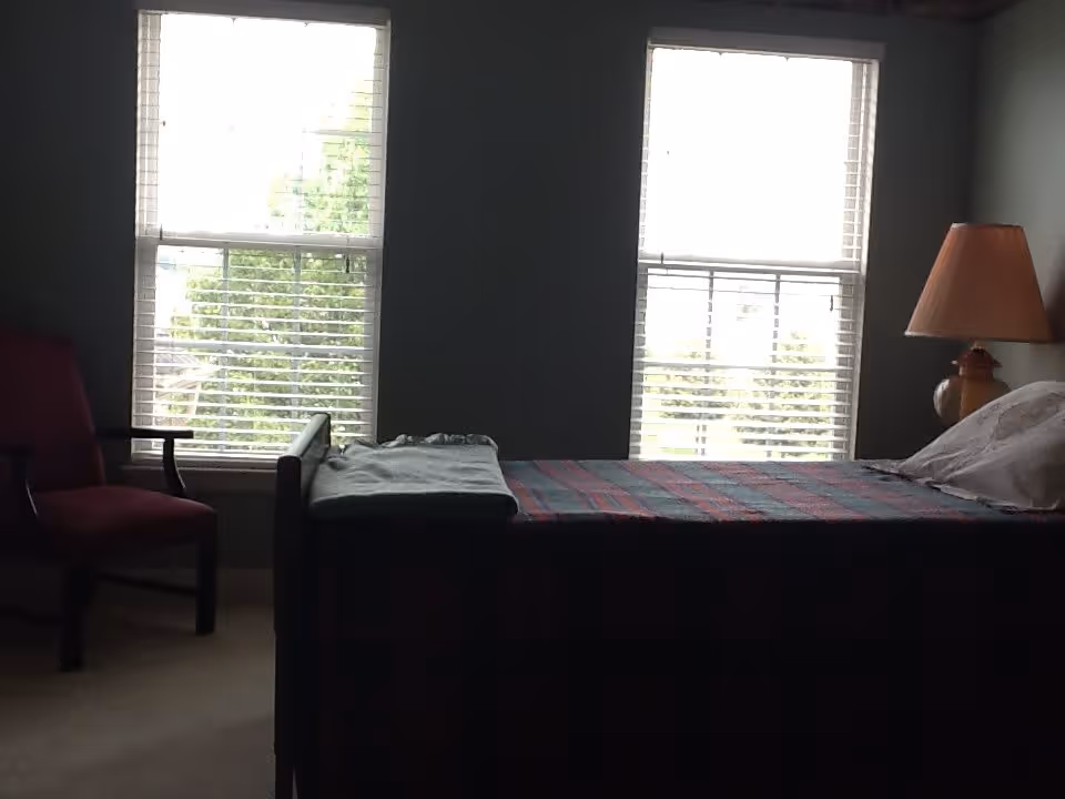 A simple bedroom with a bed, bedside lamp, armchair, and two tall windows with blinds.