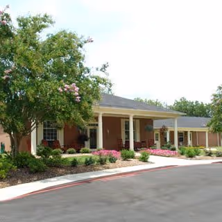 Exterior view of a single-story brick building with a covered entrance, surrounded by landscaped bushes and a tree with pink flowers, under a clear sky.