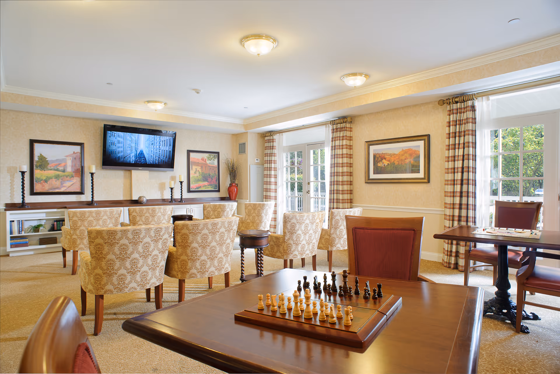 A bright and cozy senior living common area with patterned armchairs arranged facing a wall-mounted flat-screen TV. The room features framed artwork, a chessboard on a wooden table in the foreground, and large windows with plaid curtains letting in natural light.