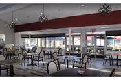 A spacious dining room with multiple tables and chairs arranged neatly. The room features large windows allowing natural light to fill the space. Modern pendant lights hang from the ceiling, and the walls are painted in neutral tones with a dark red accent beam.