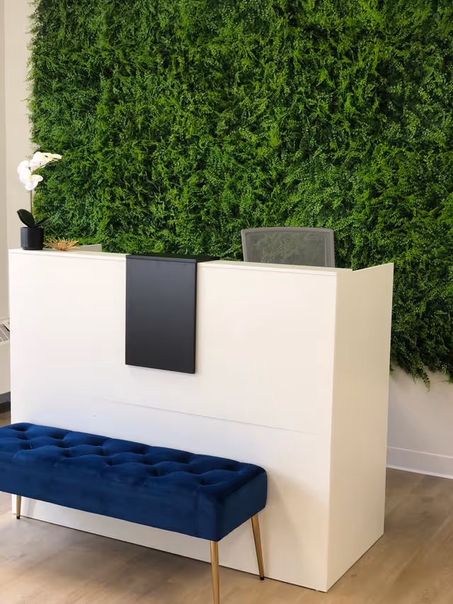 A modern reception desk with a white front panel and a black accent piece in the center. Behind the desk is a green wall covered with lush foliage. In front of the desk is a blue tufted bench with gold legs. A small potted white orchid is placed on the left side of the desk.