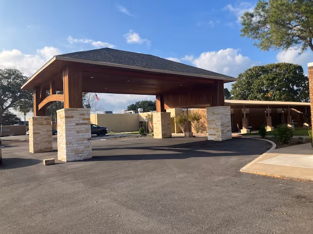 Covered driveway entrance with a wooden roof supported by stone pillars at a senior living facility, with a paved driveway and some trees and buildings in the background under a partly cloudy sky.