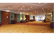 A spacious and well-lit interior hallway or lobby area with patterned carpet, recessed ceiling lights, wooden doors on the left, and a reception desk in the distance near large windows.