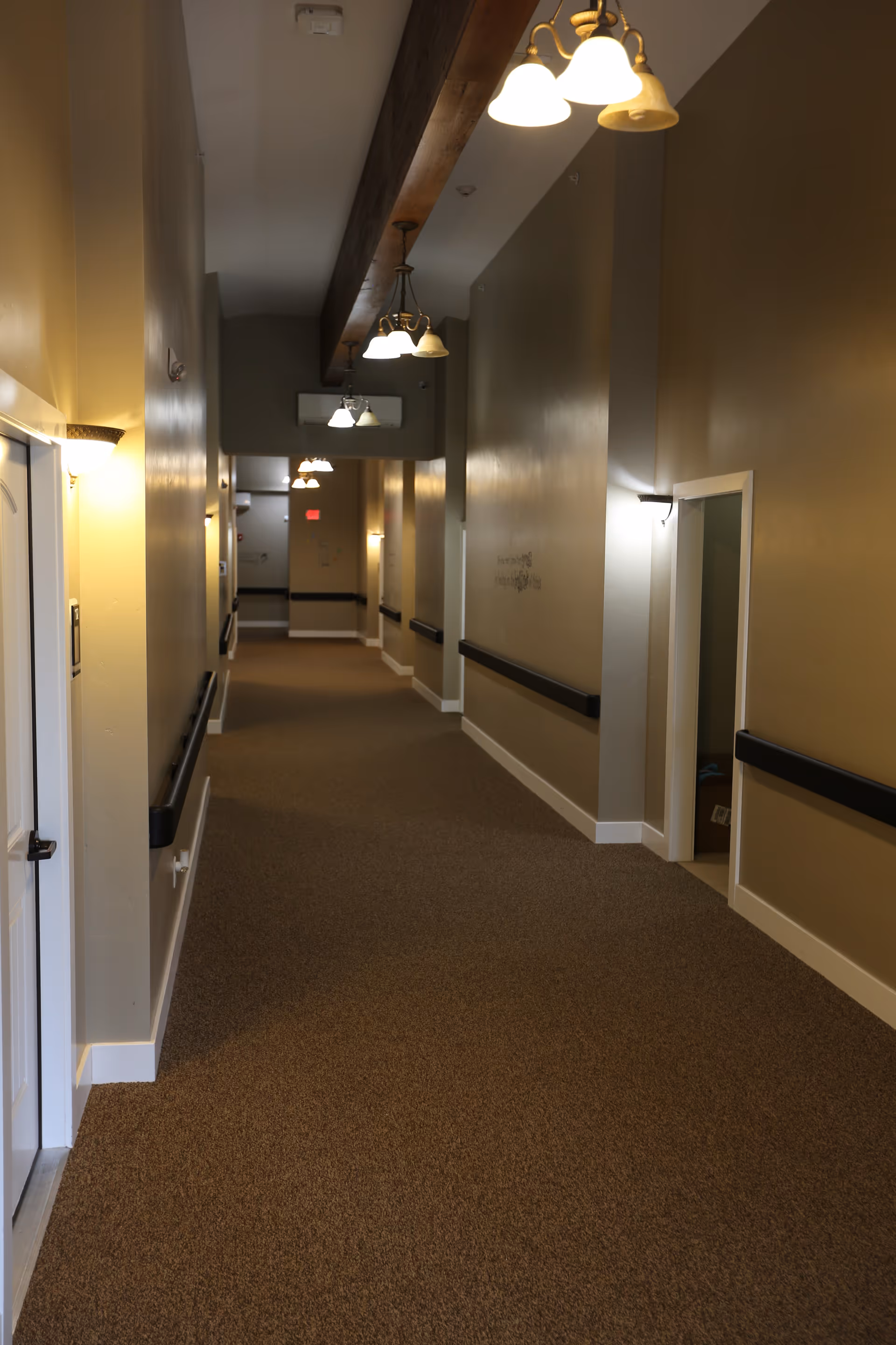 A long, carpeted hallway in a senior living facility with beige walls, white trim, and black handrails on both sides. Several ceiling light fixtures with multiple bulbs hang from a wooden beam, and wall sconces provide additional lighting. Doors and open doorways line the hallway.