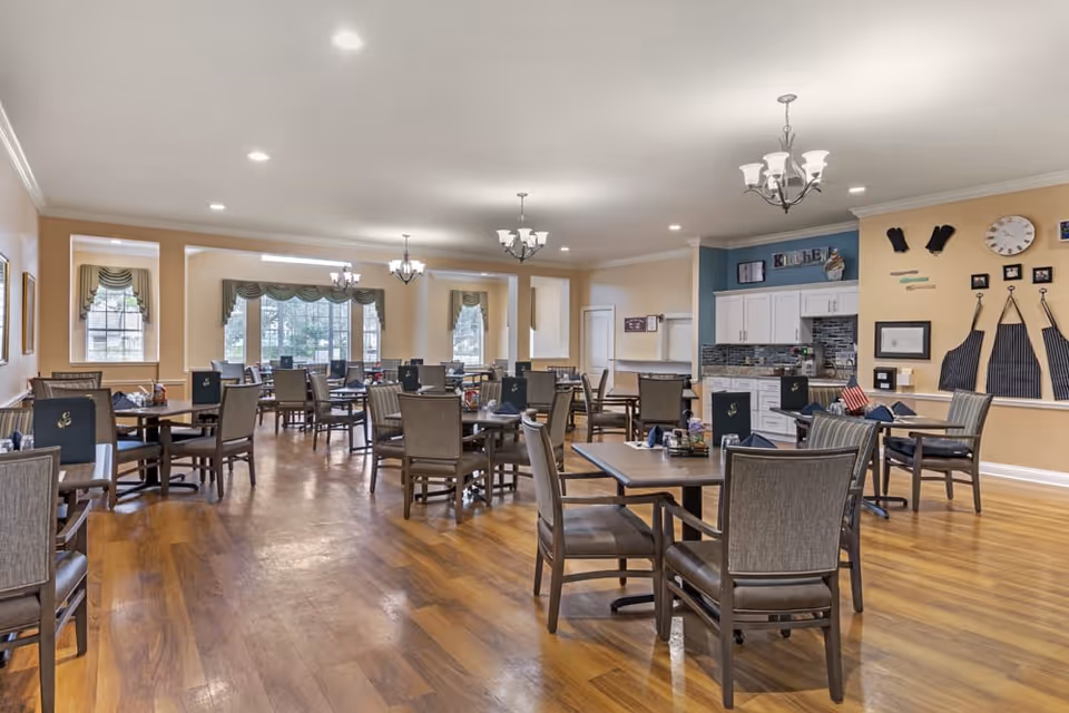 A spacious dining room with multiple tables and chairs arranged neatly on a wooden floor. The room has large windows with green valances allowing natural light to enter. There are chandeliers hanging from the ceiling providing additional lighting. On one side, there is a small kitchen area with white cabinets, a tiled backsplash, and aprons hanging on the wall. The walls are painted in warm beige tones.