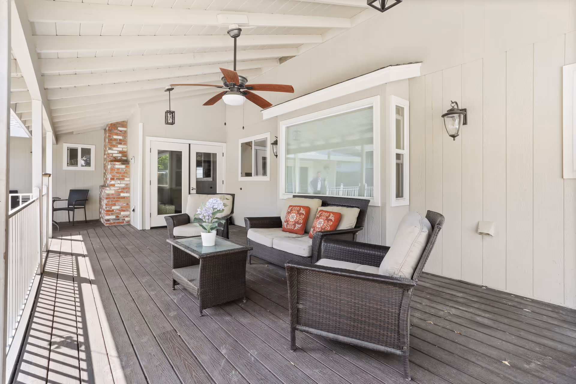 Covered outdoor patio area with wooden flooring, a ceiling fan, and wicker furniture including a loveseat, two armchairs, and a glass-top coffee table with a potted plant. The patio has white walls, a brick chimney, and large windows looking into the interior of the building.