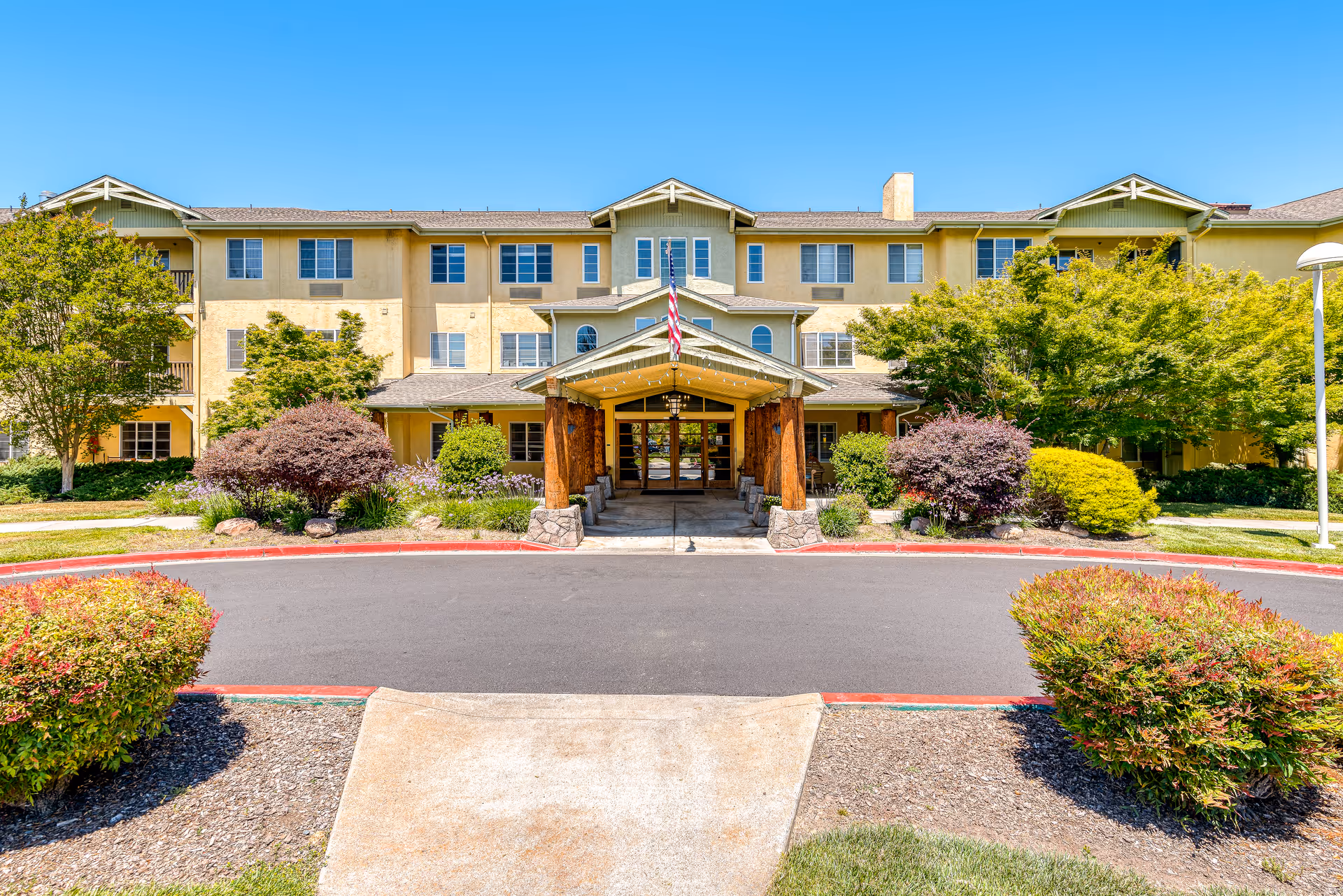 Front exterior view of a three-story senior living facility building with a covered entrance supported by wooden pillars, surrounded by well-maintained landscaping including bushes and trees under a clear blue sky.