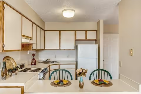 A compact kitchen area with white cabinets and countertops, a white refrigerator, and a stove with four burners. In the foreground, there is a counter with two green chairs and two place settings, each with a yellow plate and a small bunch of grapes. A small vase with dried flowers is placed between the two settings. The walls are beige, and there is a ceiling light fixture above.