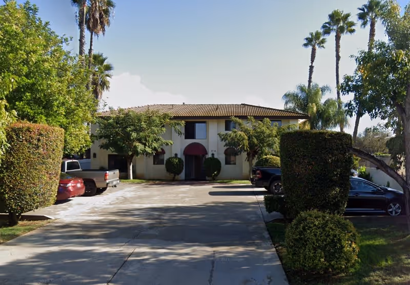 Front exterior view of a two-story building with a tiled roof, surrounded by trimmed bushes and palm trees. There is a driveway leading up to the entrance, with several parked cars on either side.