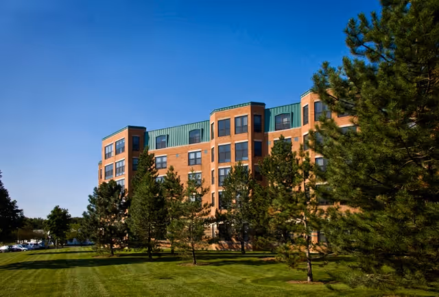Brick multi-story residential building with a green roof, trees, and a manicured lawn under a clear blue sky.