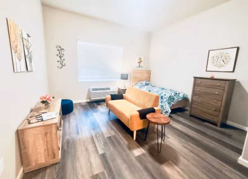 A bright senior living bedroom with a single bed covered in a blue and white patterned blanket, a mustard yellow loveseat with black armrest covers, a small round wooden side table, a wooden dresser, a wooden cabinet with flowers and magazines on top, a blue pouf, and a window with blinds letting in natural light.