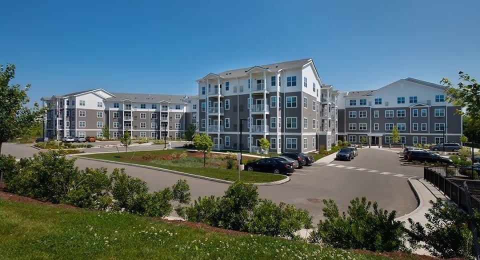 Exterior view of a large, modern senior living facility with multiple connected buildings, parking spaces with cars, landscaped greenery, and clear blue sky.