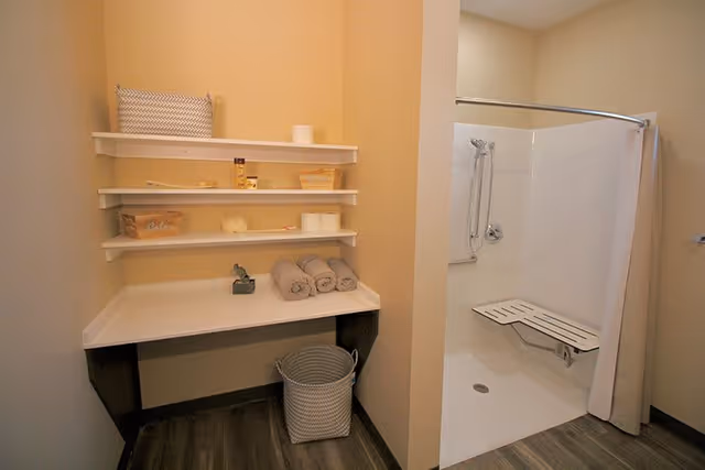 A bathroom area with a walk-in shower featuring a foldable bench and grab bars. Next to the shower is a countertop with three rolled towels, a small decorative item, and shelves above holding various toiletries and toilet paper rolls. A woven basket is placed under the countertop.