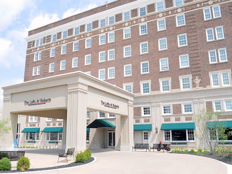 Exterior view of The Lofts at Roberts, a multi-story brick building with numerous windows, a covered entrance with the facility name displayed, green awnings over windows, benches, and some landscaping including small trees and bushes.