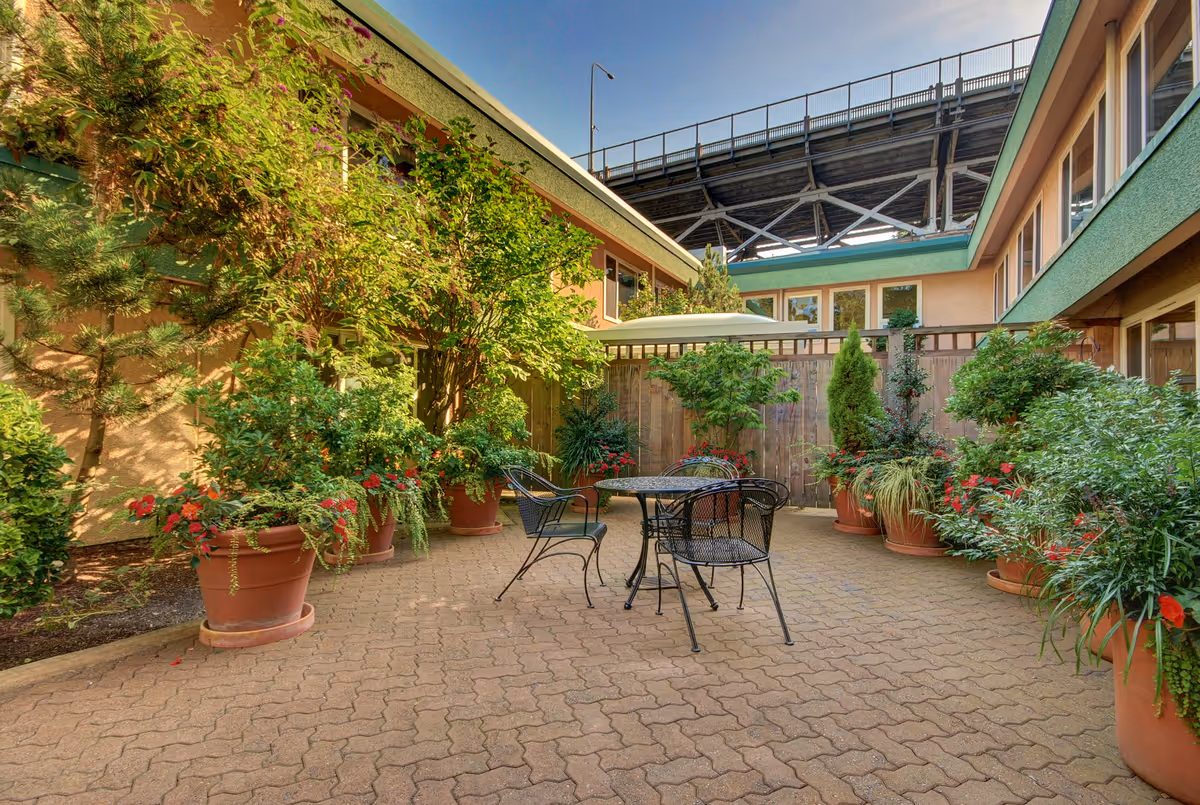 Outdoor courtyard area with potted plants and greenery surrounding a metal table and four chairs. The courtyard is enclosed by the building walls and a wooden fence, with a large bridge structure visible in the background under a clear blue sky.