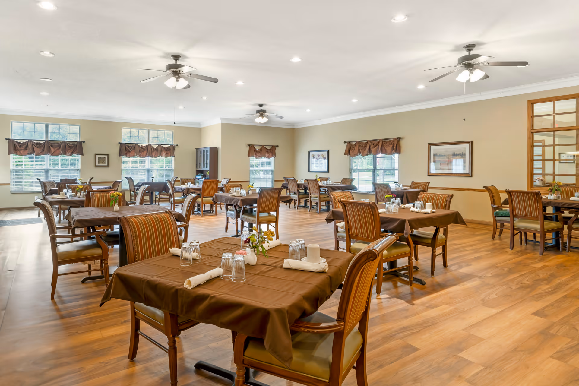 A spacious dining room with multiple tables covered in brown tablecloths, each set with glasses, napkins, and small flower vases. The room has wooden floors, several windows with brown valances, ceiling fans with lights, and framed artwork on the walls.