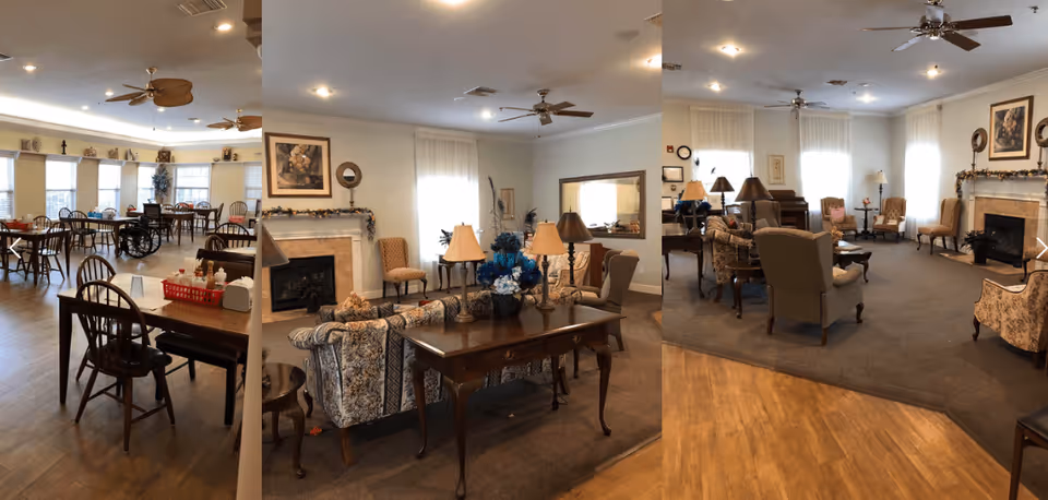 A spacious and well-lit assisted living facility interior featuring a dining area with multiple wooden tables and chairs, some with red baskets and condiments on top. Adjacent to the dining area is a cozy living room with floral patterned sofas, armchairs, a wooden coffee table, lamps, and a fireplace decorated with garlands. The room has ceiling fans, large windows with sheer curtains, and framed artwork on the walls.
