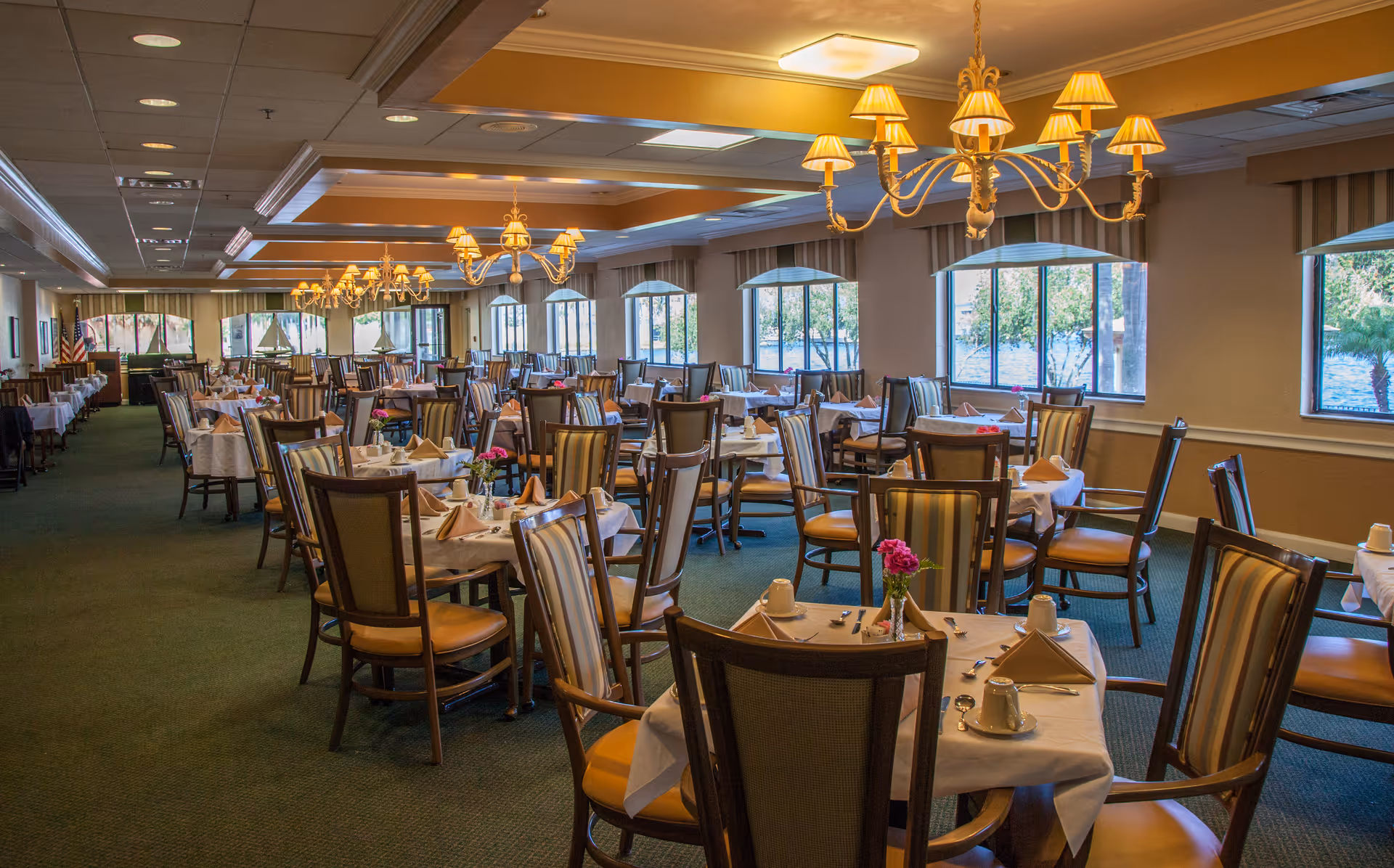 Large dining room with multiple tables set for service, upholstered chairs, chandeliers, and windows along one wall.