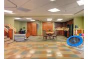 A spacious room with a tiled floor featuring a checkered pattern in pastel colors. The room includes a small dining table with chairs, a kitchenette area with wooden cabinets, and a staircase on the left side. There is a blue trampoline on the right side and a cushioned bench near the stairs. The ceiling has recessed lighting panels.