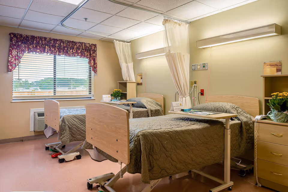 A bright twin patient room with two adjustable beds, over-bed tables, bedside furniture and a window with a floral valance.