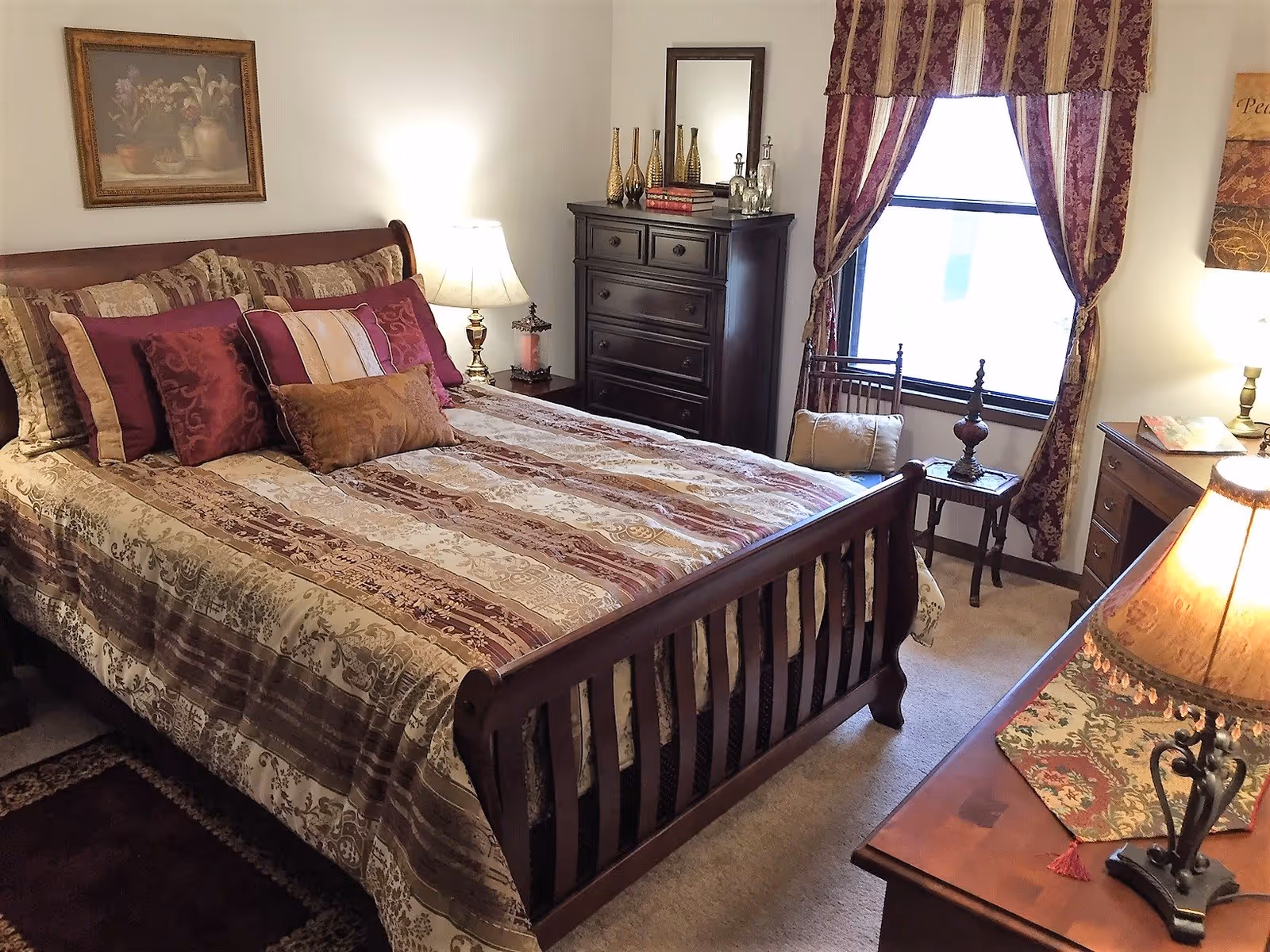 A cozy bedroom with a wooden bed frame and a bed made with patterned bedding and multiple decorative pillows in shades of burgundy, gold, and beige. There is a wooden dresser with decorative items and a mirror on top, a window with burgundy and gold curtains, a small side table with a lamp and cushion, and a wooden desk with a lamp and a decorative runner.