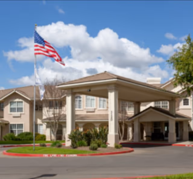 Exterior view of Marbella Visalia senior living facility showing the main entrance with a covered driveway, an American flag on a flagpole, and a clear blue sky with some clouds.