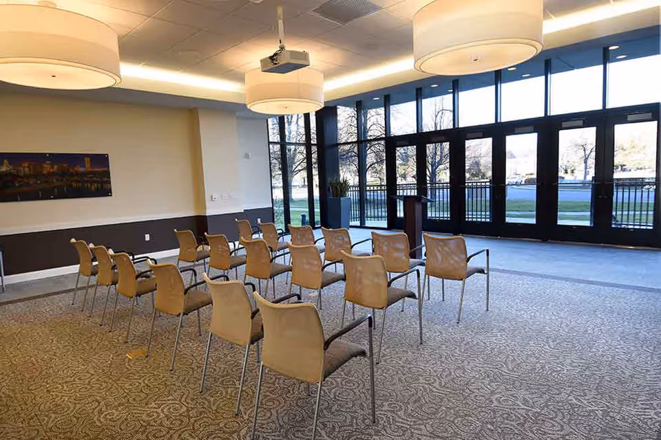 A bright multipurpose meeting room with rows of chairs facing a podium and large floor-to-ceiling windows.