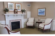 A cozy sitting area with a fireplace, three upholstered armchairs, a small side table with a decorative item, and framed artwork on the walls. The room has neutral tones and a carpeted floor.
