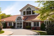 Front exterior of a brick-and-siding senior living facility with a rounded glass entrance, arched windows, and a circular driveway.