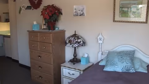 A cozy bedroom with a wooden dresser topped with a poinsettia plant, tissue box, and framed photo. Next to the dresser is a nightstand with a decorative lamp and another tissue box. The bed has a purple blanket and blue floral pillows. The walls are decorated with a black and white photo and a framed painting.