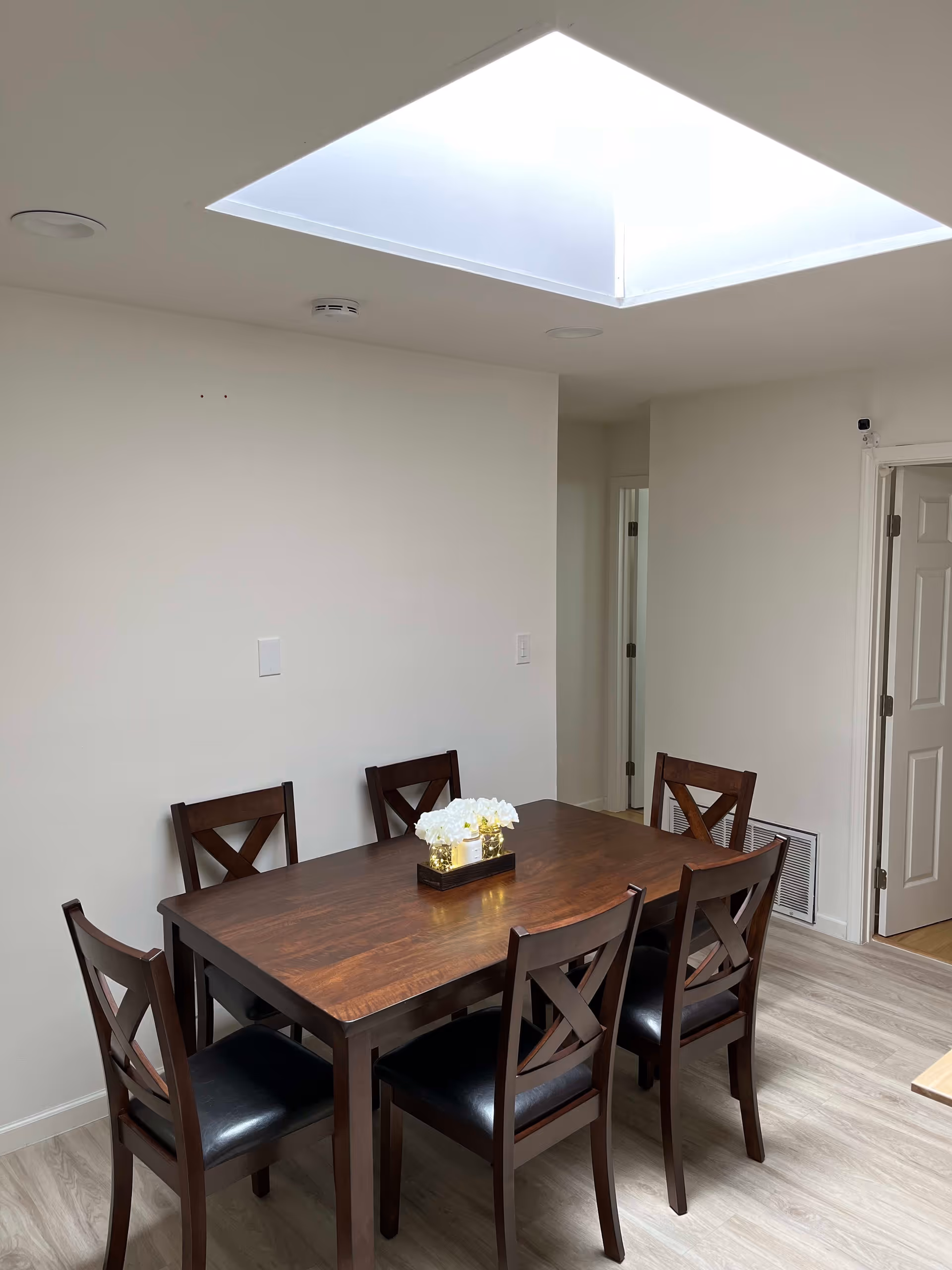 A dining area with a wooden table and six matching chairs with black cushions. A rectangular tray with white flowers and small decorative lights is placed in the center of the table. The room has light-colored walls and flooring, and a large skylight on the ceiling providing natural light. There are two doorways visible in the background.