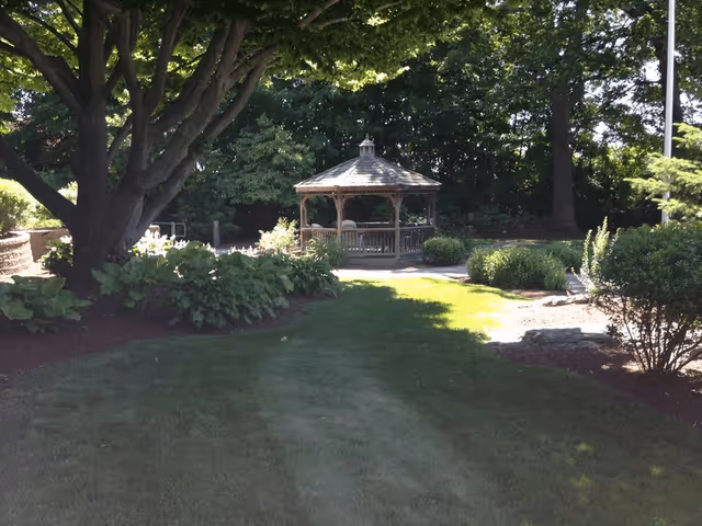 Wooden gazebo centered in a shaded garden with large trees, shrubs, and a grassy lawn.