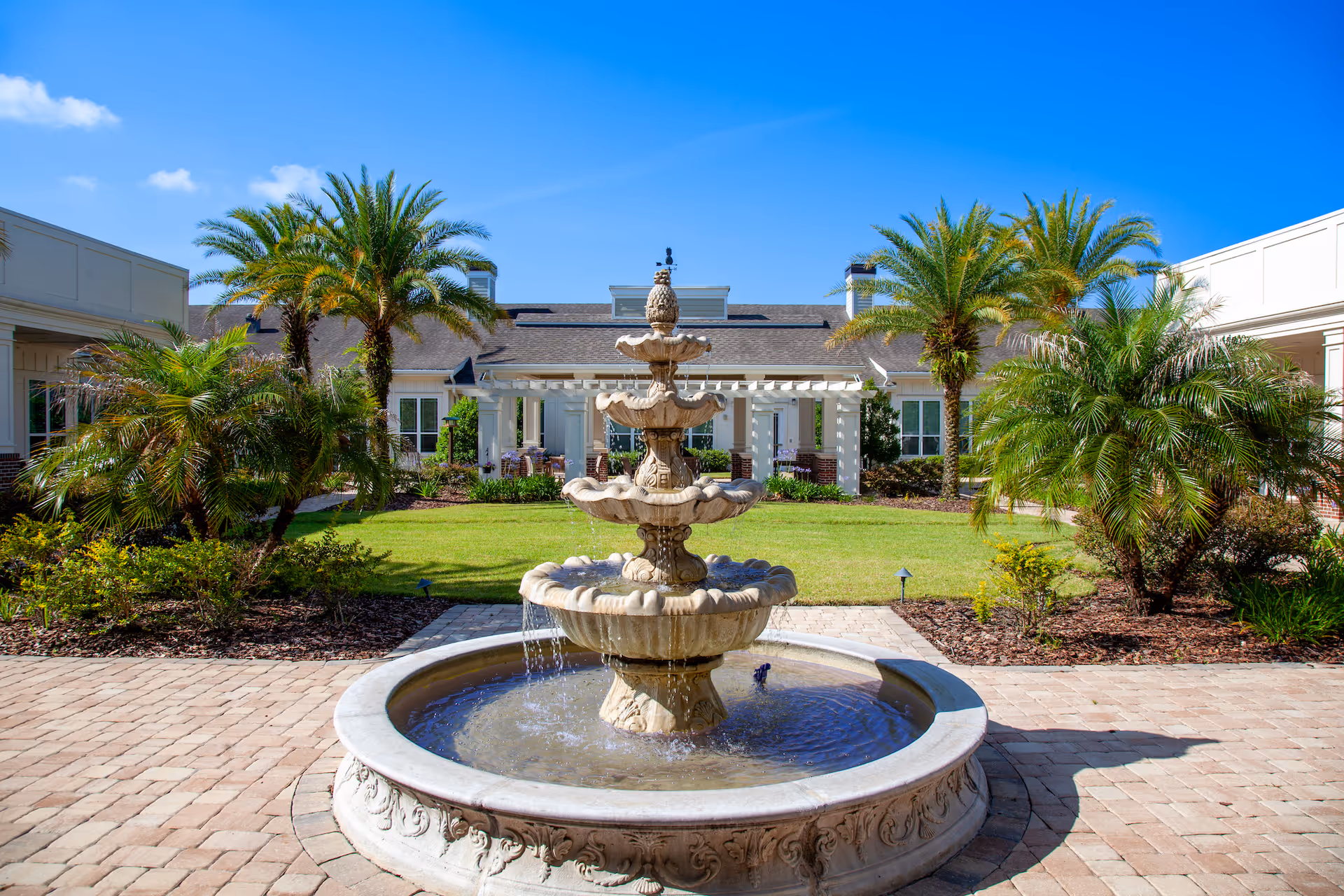 Outdoor courtyard area at San Jose Gardens featuring a multi-tiered stone fountain in the center, surrounded by palm trees, green grass, and a paved walkway. The building with white walls and windows is visible in the background under a clear blue sky.