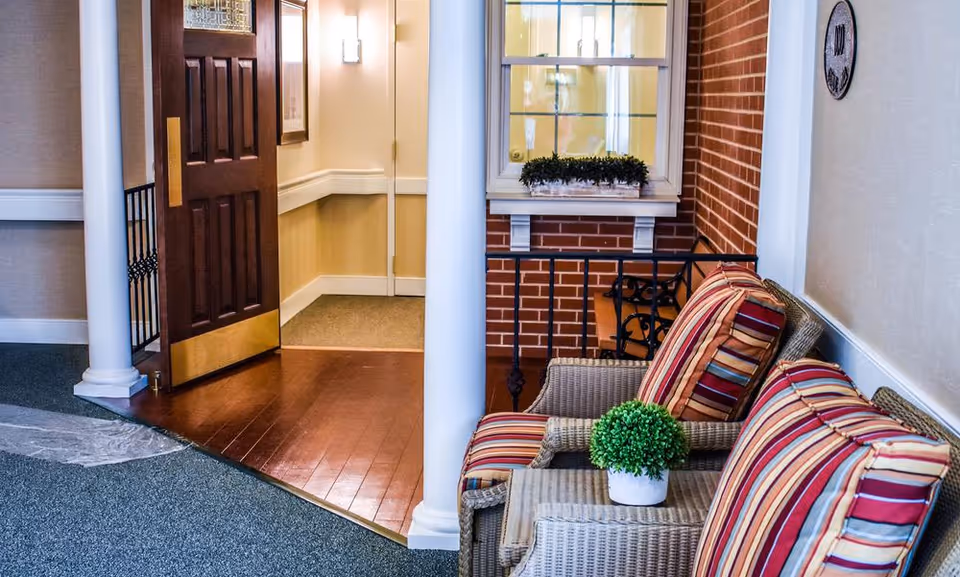 A cozy indoor seating area with two wicker chairs featuring striped cushions in red, beige, and green colors. A small table between the chairs holds a small green potted plant. The area has a carpeted floor transitioning to wooden flooring near an open dark wooden door with a brass kick plate. The walls are light-colored with white trim, and there are white columns and a window with a small plant box on a brick wall in the background.