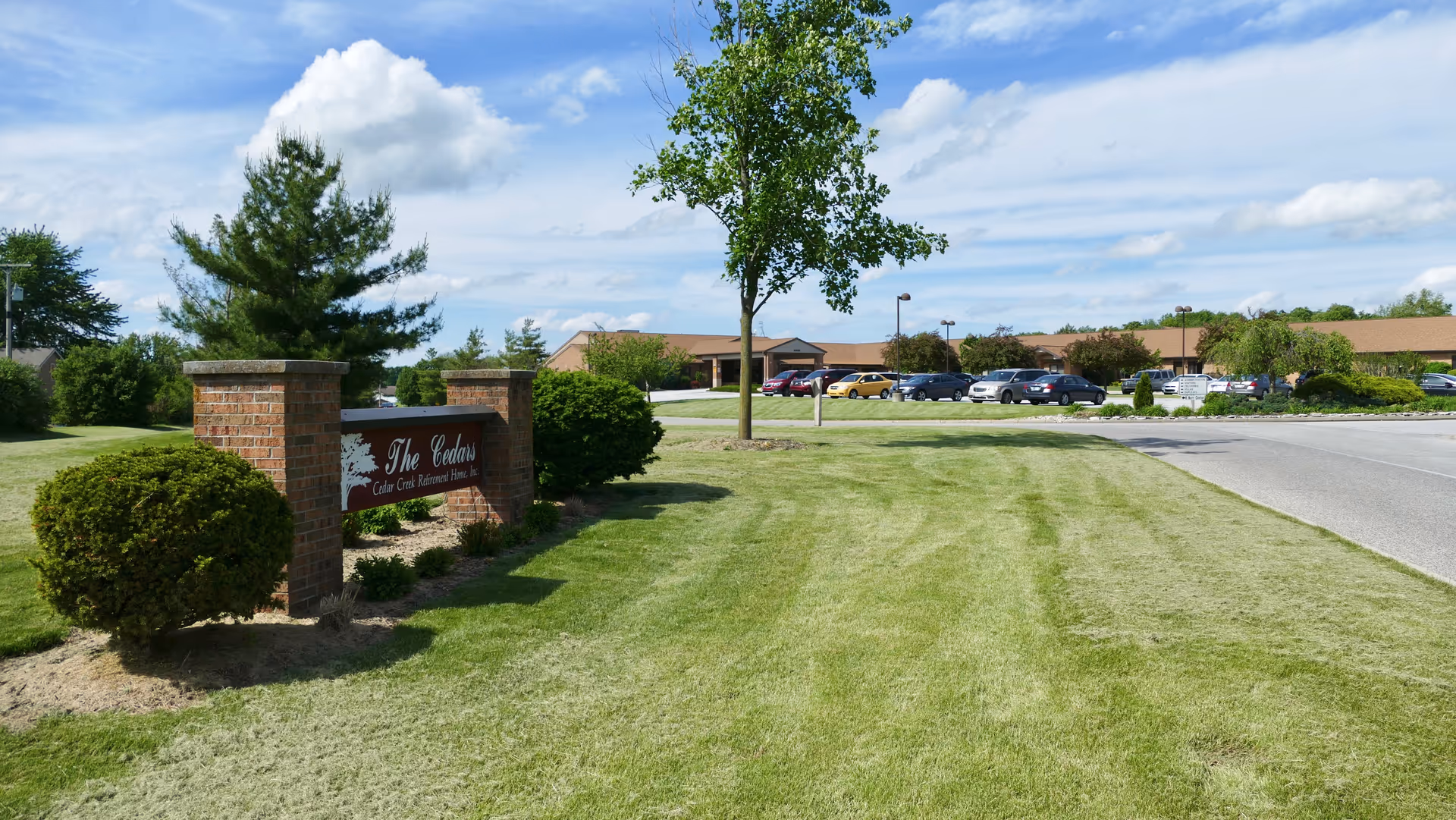 Outdoor view of The Cedars senior living facility with a brick sign displaying the facility name, well-maintained green lawn, trees, bushes, a parking lot with several cars, and a partly cloudy sky.