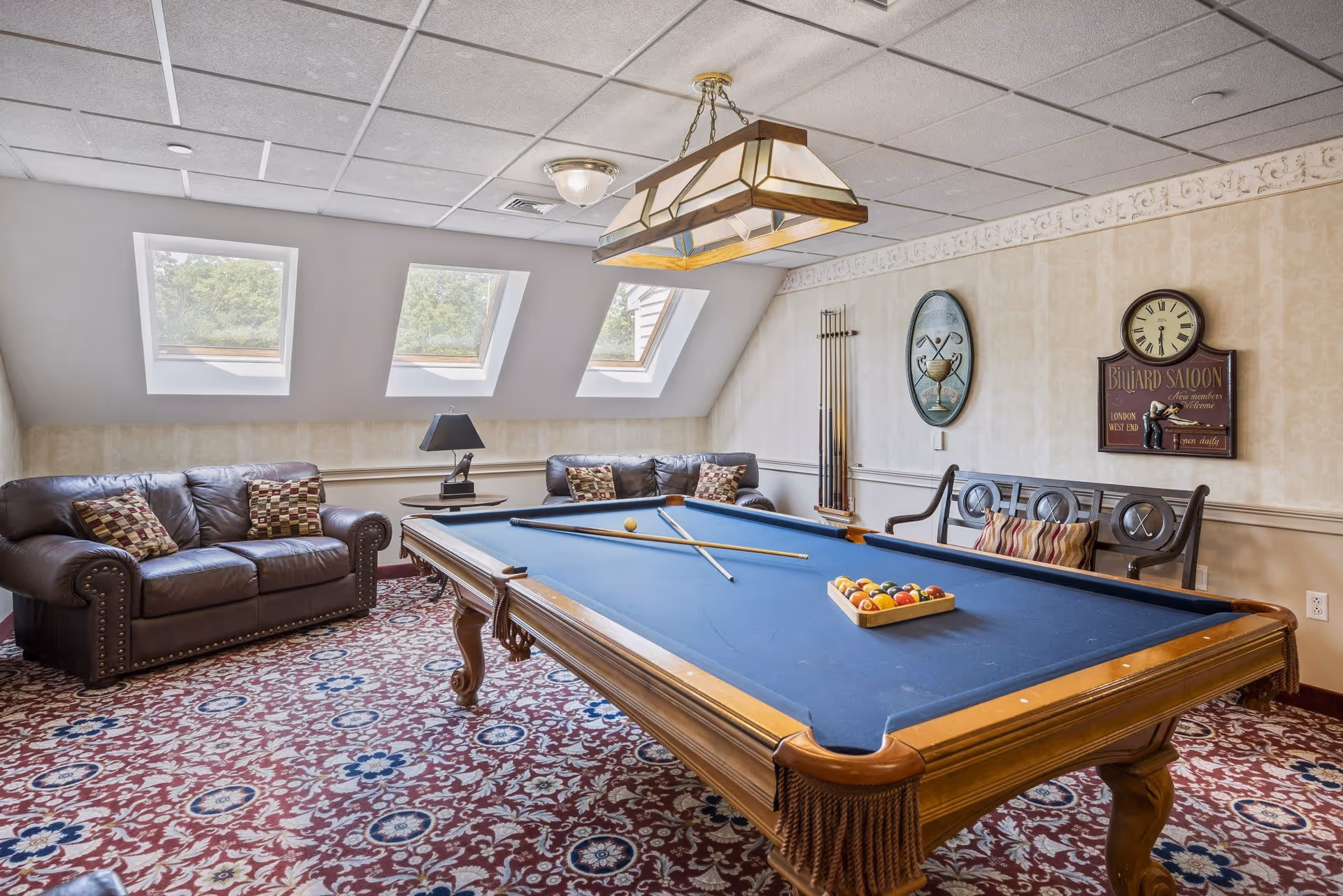 A cozy game room with a blue felt pool table in the center, two leather sofas with patterned cushions, a decorative lamp hanging above the pool table, three windows letting in natural light, a wall clock, and billiard cues mounted on the wall.