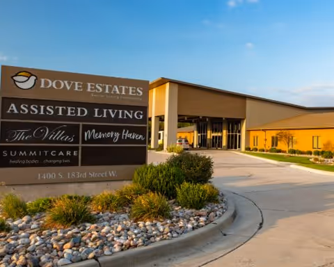Exterior view of Dove Estates Senior Living Community building with a large sign in the foreground displaying services like Assisted Living, The Villas, Memory Haven, and Summit Care. The building is surrounded by landscaping with bushes and rocks, and a driveway curves around the sign.