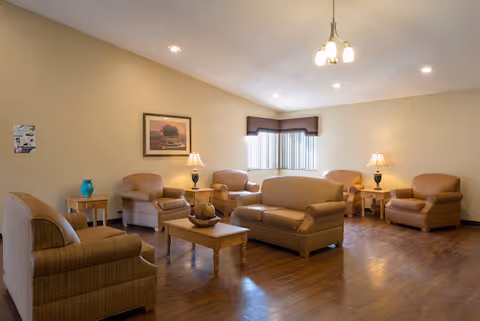A well-lit living room with beige walls and wooden flooring featuring a sofa, several armchairs, two side tables with lamps, a coffee table with decorative items, a framed picture on the wall, and a window with vertical blinds and a valance.