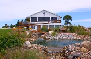 Two-story white building with a wooden balcony, landscaped rocks, and a pond in front.
