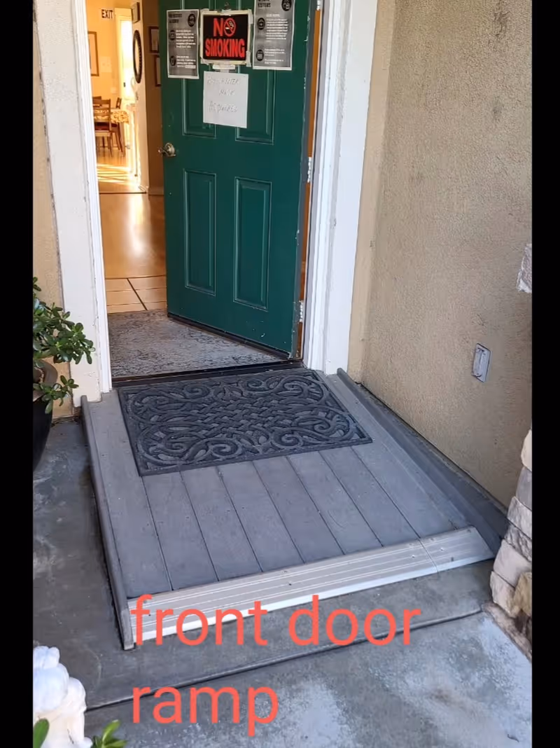 Open green front door with a decorative black doormat on a gray ramp leading up to the entrance. The door has several signs including a 'No Smoking' sign and a handwritten note. The interior of the building is partially visible, showing a tiled floor and some furniture. The exterior wall is beige with a small plant visible on the left side.